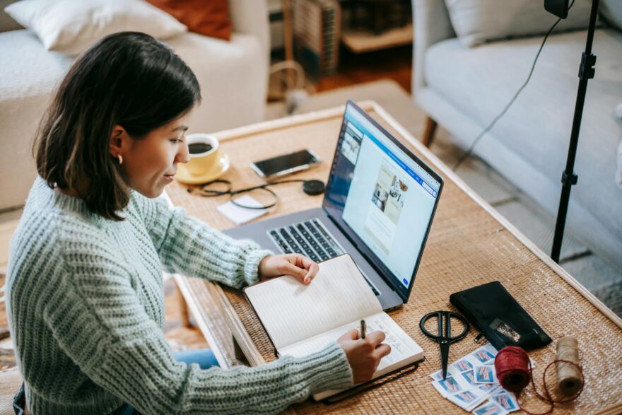 young focused woman writing in planner while using laptop